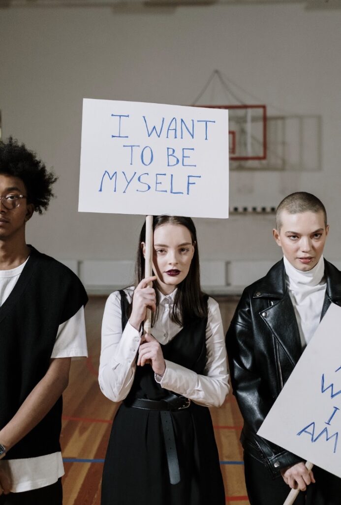 Person wearing dark clothes and dark lipstick carrying a sign that says "I want to be myself." They are standing in between two other people.