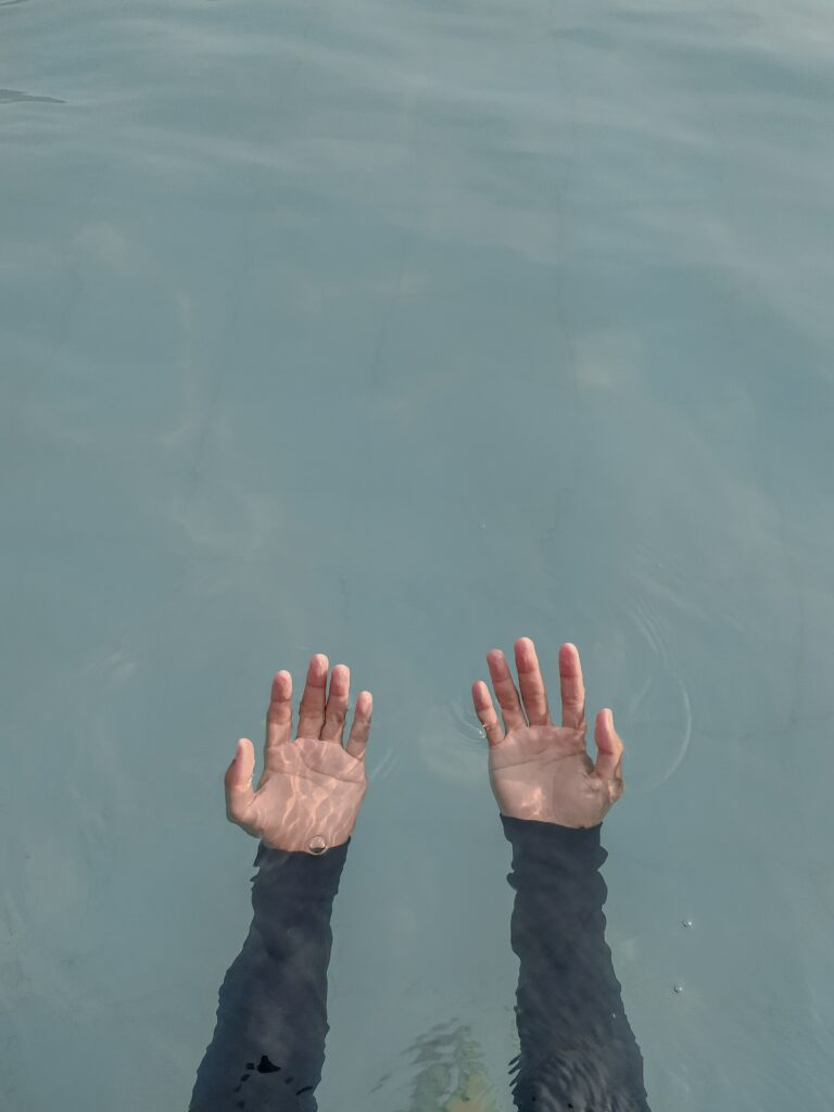 Two outstretched arms, show from the elbow to the fingertips, palms upward. They are submerged underwater, with the fingertips just out of the water. The water is bluish and clear, and sunlight reflects on it. This is a way that you could feel emotionally numb.